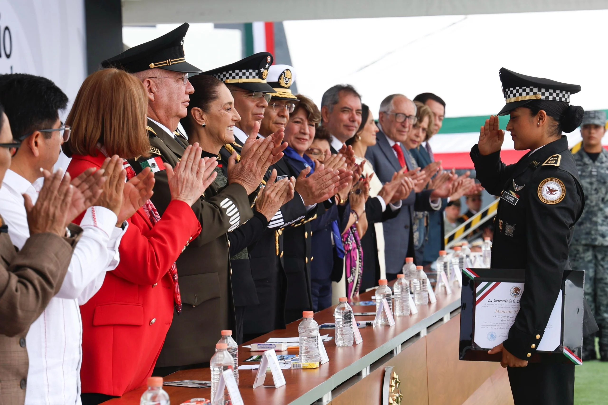 Ficha de imagen Texto alternativo (alt): Presidenta Claudia Sheinbaum y funcionarios aplauden a una elemento de la Guardia Nacional que recibe un reconocimiento durante el 7° aniversario de la institución.