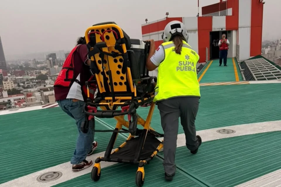 Personal de SUMA Puebla traslada camilla de emergencia en helipuerto sobre azotea, con vista de la ciudad al fondo.